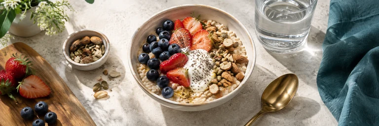 Overhead appetizing balanced bowl of oats, berries, yogurt and nuts beside a clear water glass, light stone counter and teal linen, real food textures.